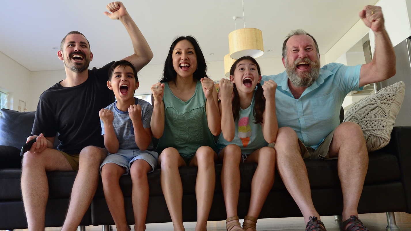 Family of five sitting on a couch in a living room, with two adults and three children cheering.